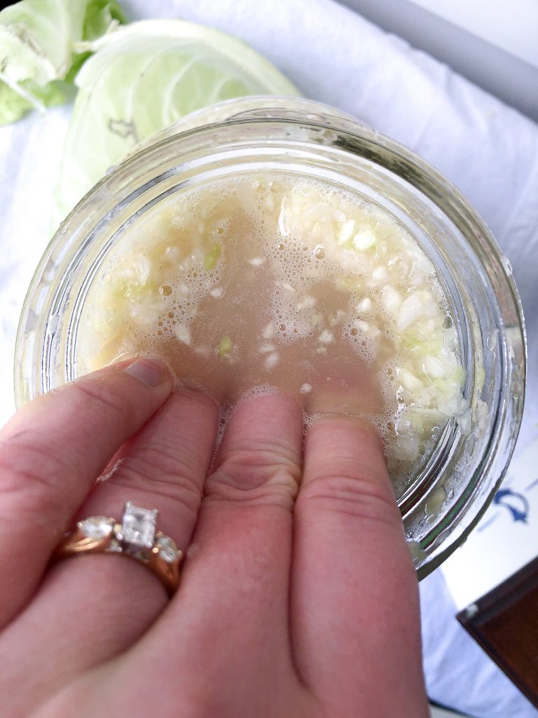 hand pushing cabbage leaves down into a jar of sauerkraut with brine covering fingers.
