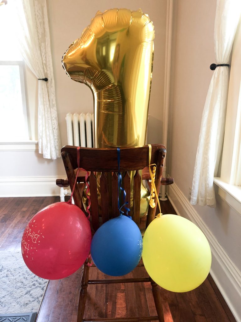 antique wooden high-chair with red, blue and yellow balloons tied on the back, and a big gold number 1 balloon.