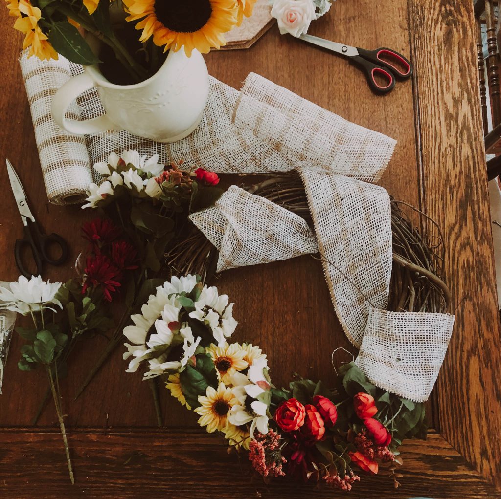 seasonal wreath making materials laid out on wood table and a wreath sitting next to a pitcher of sunflowers