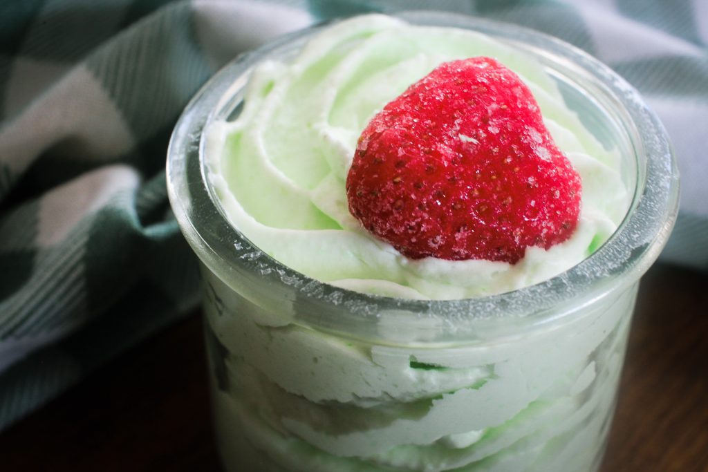 close up of mousse in a glass jar with a strawberry on top in front of a green gingham towel